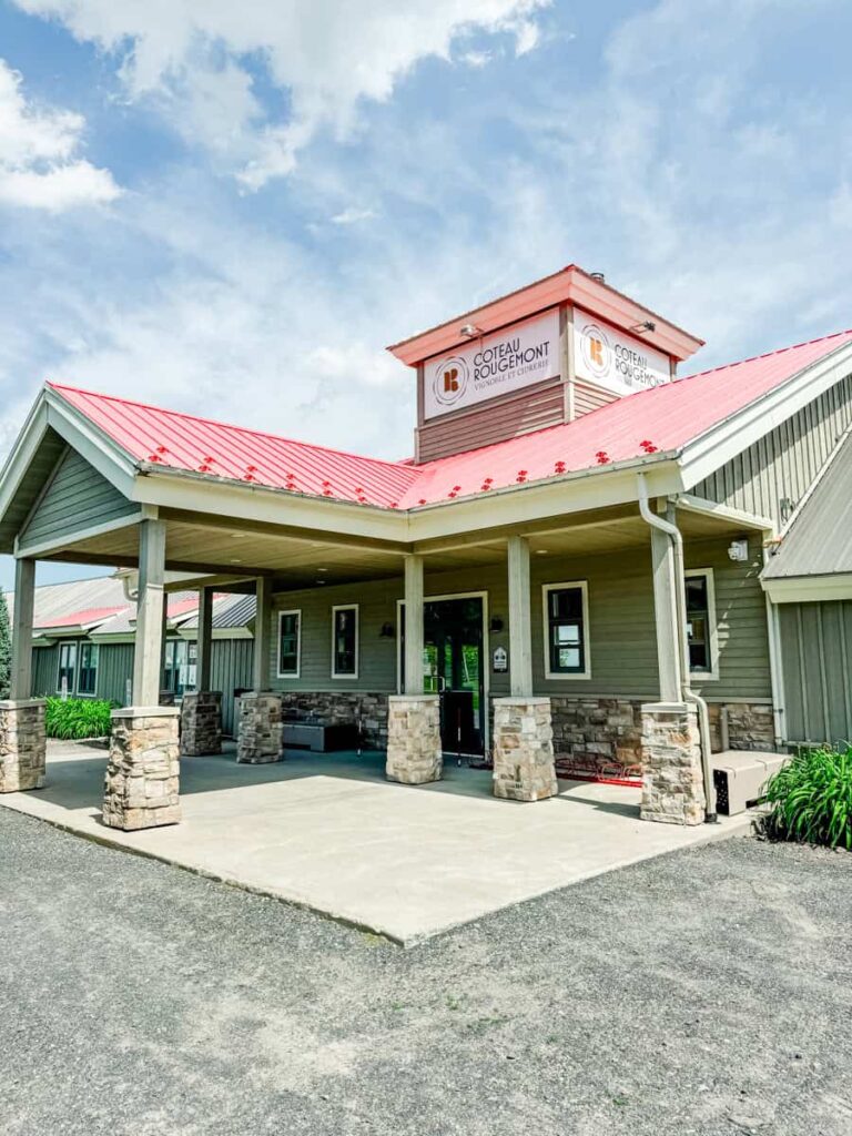 Exterior of the Coteau Rougemont winery, a modern barn-style building with a red metal roof and stone pillars under a bright blue sky.