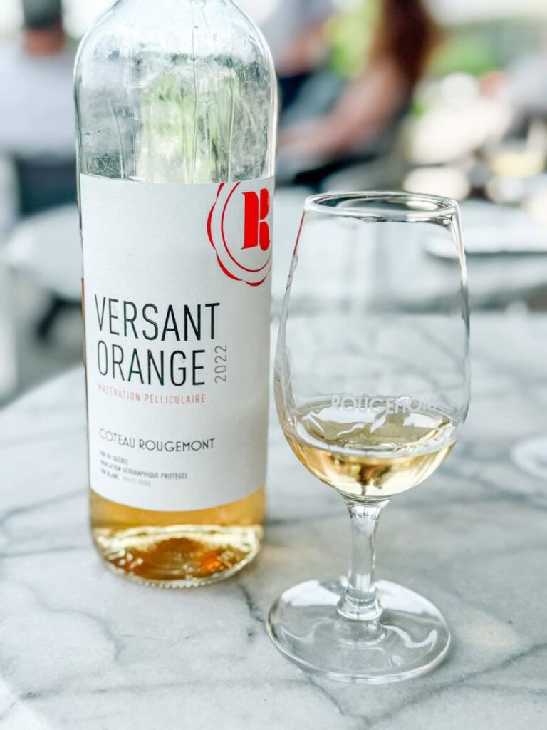 Close-up of a chilled bottle of Versant Orange 2022 white wine from Coteau Rougemont beside a half-filled branded wine glass on a marble tabletop.