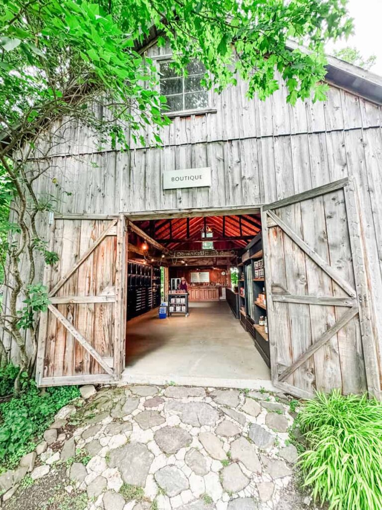 Weathered wood barn at Domaine du Ridge with its double doors open, inviting guests into a boutique surrounded by greenery and a stone path.