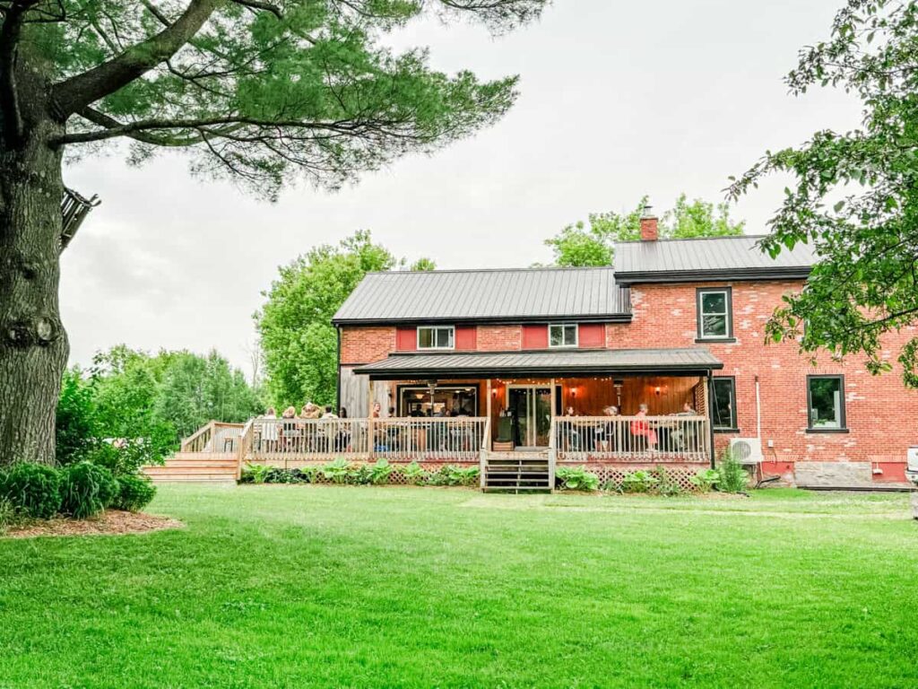 Exterior view of Espace Old Mill, a red brick farmhouse-style restaurant with a covered porch and patio dining area, surrounded by green lawn and trees.