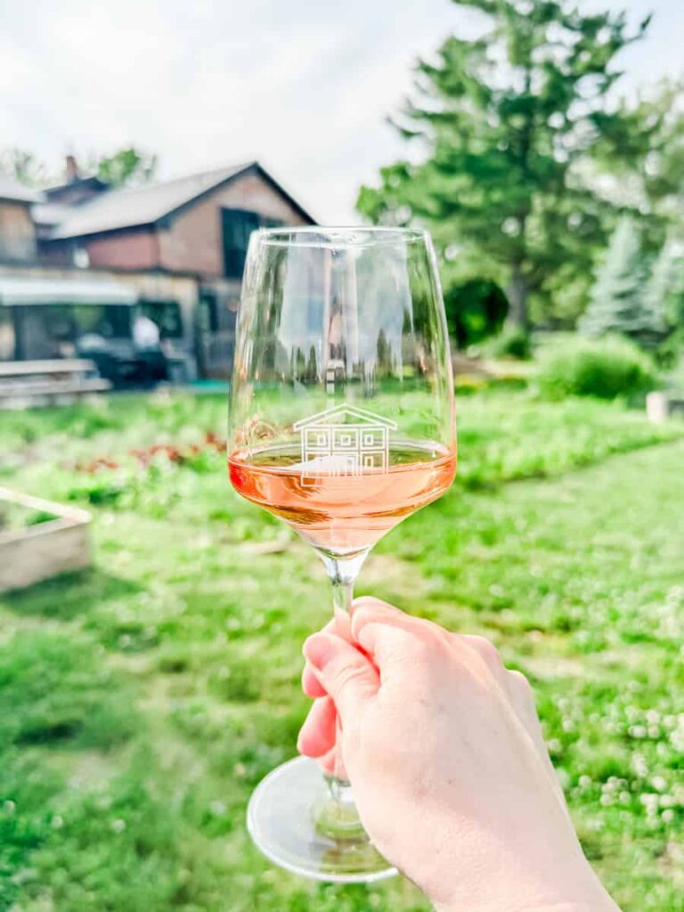 Hand holding a glass of rosé wine with the Espace Old Mill logo, overlooking garden beds and a grassy outdoor dining area.