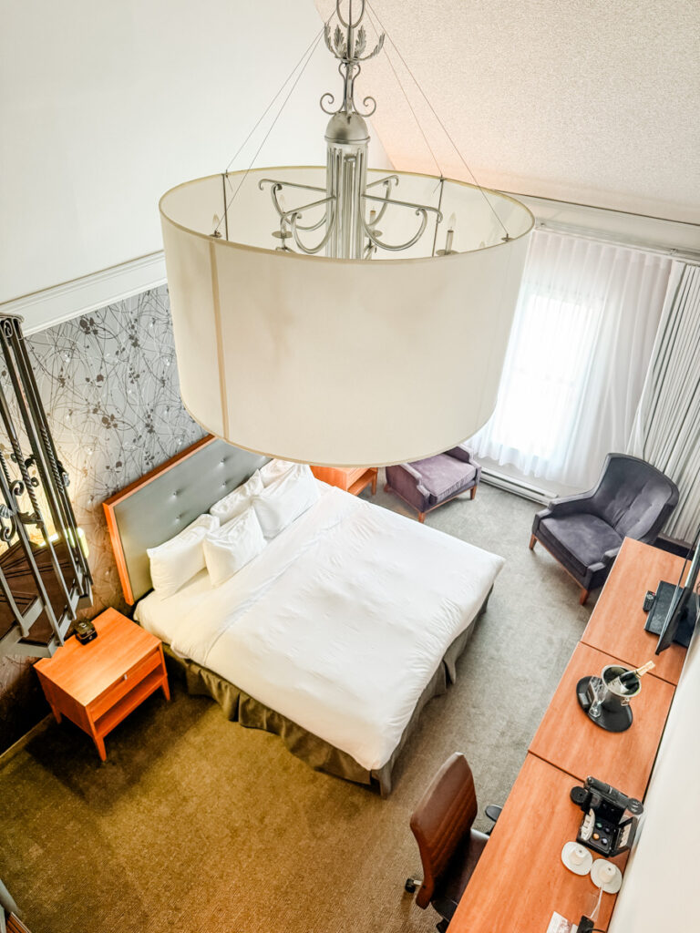 Overhead view of a cozy hotel room featuring a queen bed, two armchairs, a large desk, and a chandelier, with soft natural light from a curtained window.