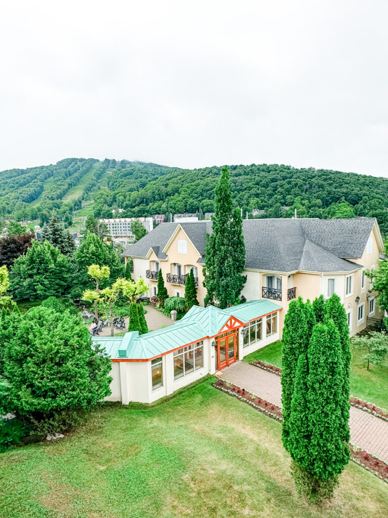 Aerial view of the Château Bromont property, showing a pale yellow hotel building with a green roof and manicured landscaping, set against the lush backdrop of Mont Brome.