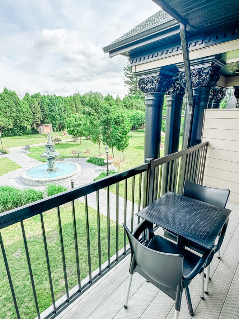A private balcony at Hotel Montfort with a black patio table and chairs overlooks a landscaped garden with a fountain and walking paths.