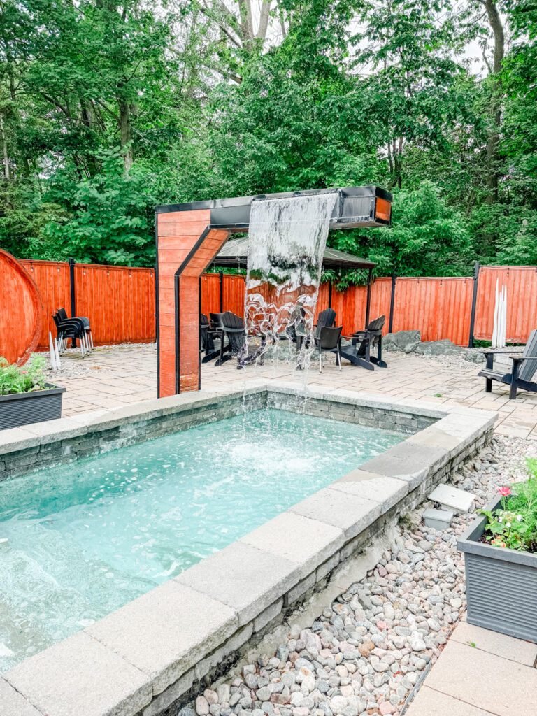An outdoor spa area at Hôtel Montfort with a stone-lined cold plunge and cascading waterfall, surrounded by trees and wooden fencing for privacy.