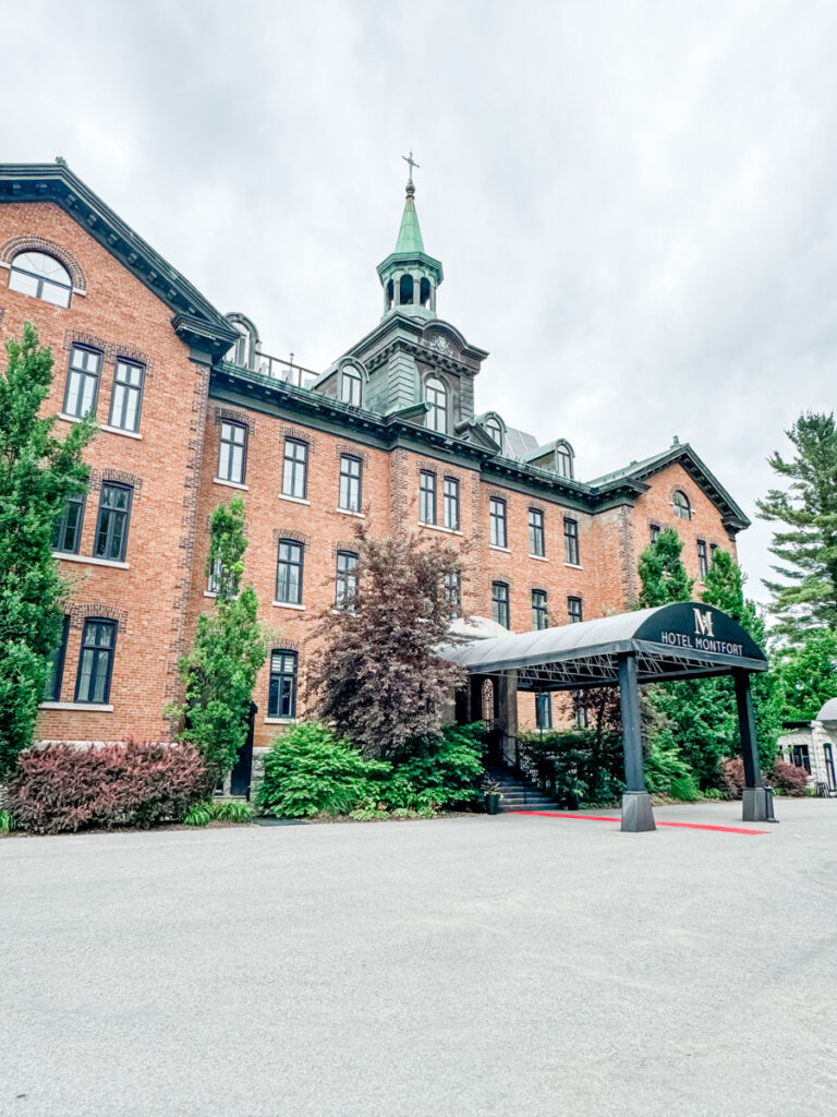 The historic brick exterior of Hotel Montfort, topped with a green steeple, with a black awning at the entrance and greenery along the walkway.