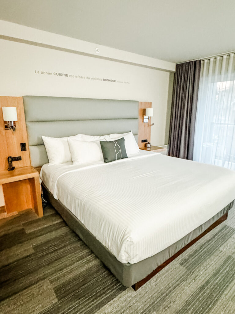A modern guest room at Hotel Rive-Gauche featuring a king bed with a soft gray headboard, wood-accented nightstands, and a quote on the wall in French about cuisine and happiness.