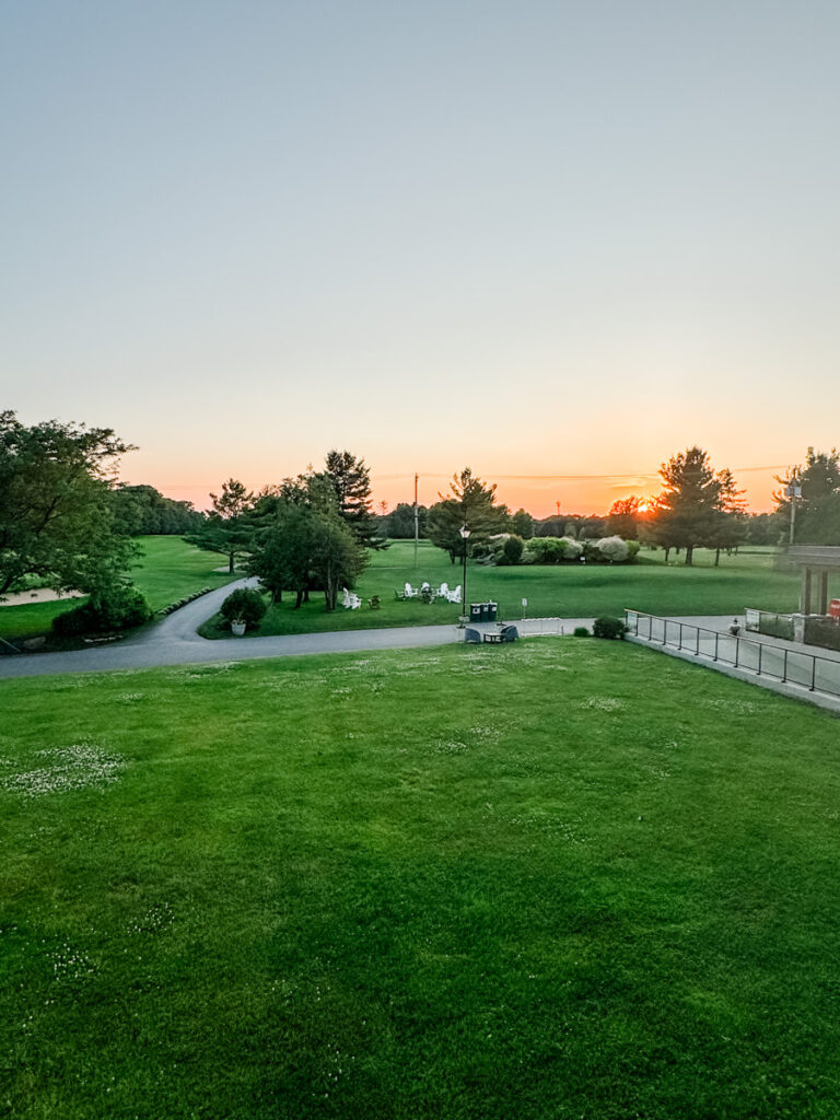 A wide view of the green golf course at La Cache du Lac Champlain, with a tree-lined path and Adirondack chairs in the distance, captured during a serene sunset.