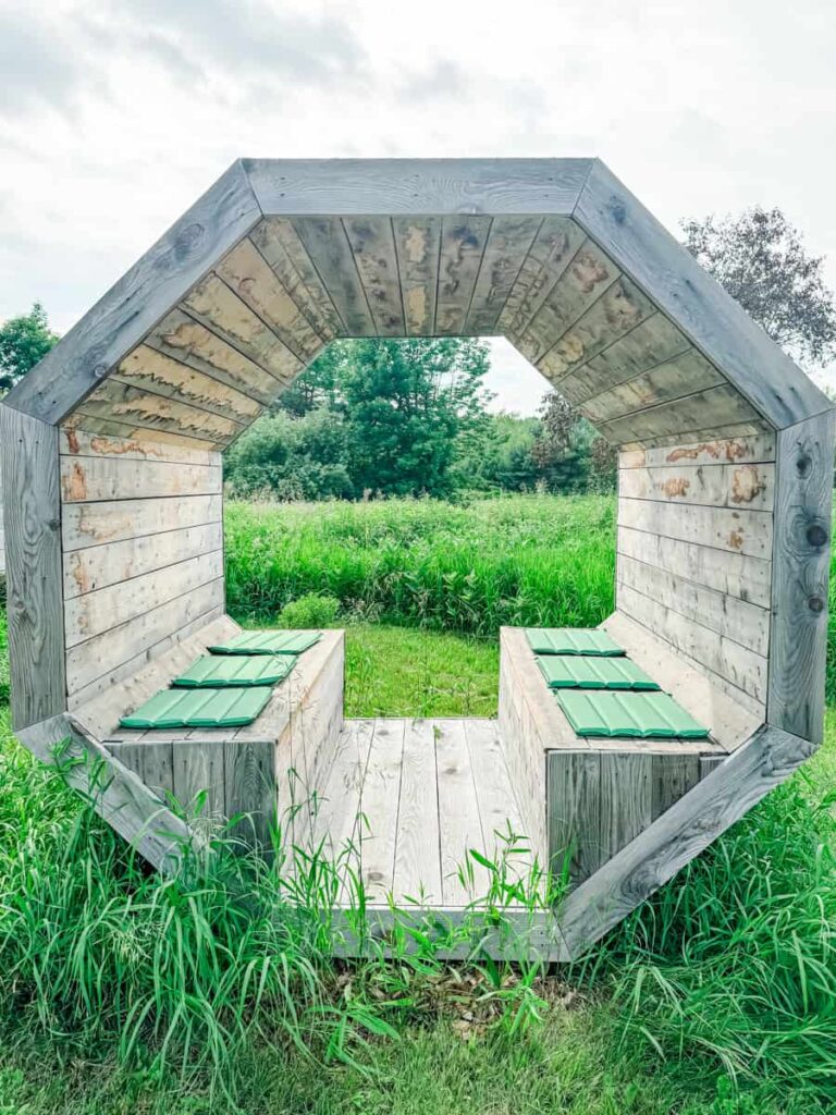 Octagonal wooden structure with green-seated benches set in tall grass, part of the outdoor setting at Les Cocagnes.