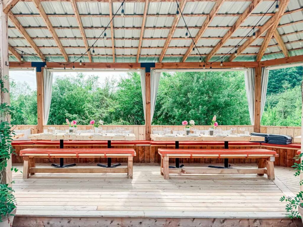 Covered outdoor dining area at Les Cocagnes with long wooden tables, benches, string lights, and wild greenery in the background.