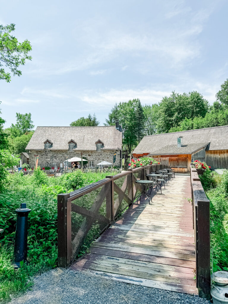 A wooden bridge leads to Moulin Michel, a historic stone mill surrounded by lush greenery, with patio tables and umbrellas set up for outdoor dining.