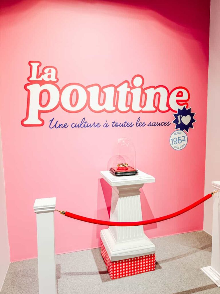 A bright pink museum wall displays the title La Poutine with the tagline “Une culture à toutes les sauces,” behind a velvet rope and pedestal featuring a poutine sculpture under glass.