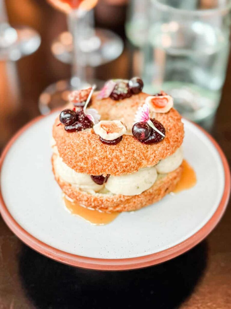 Paris-Brest maple crémeux dessert, filled with creamy maple and blackcurrant layers, topped with northern hazelnuts and edible flowers, plated on a white dish.