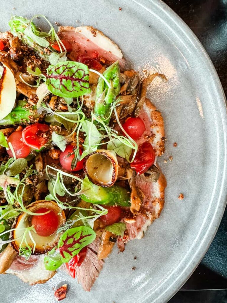 Plated smoked duck with a salad of eggplant, black garlic caviar, local asparagus, mushrooms, and sour cherries, garnished with fresh herbs and greens.
