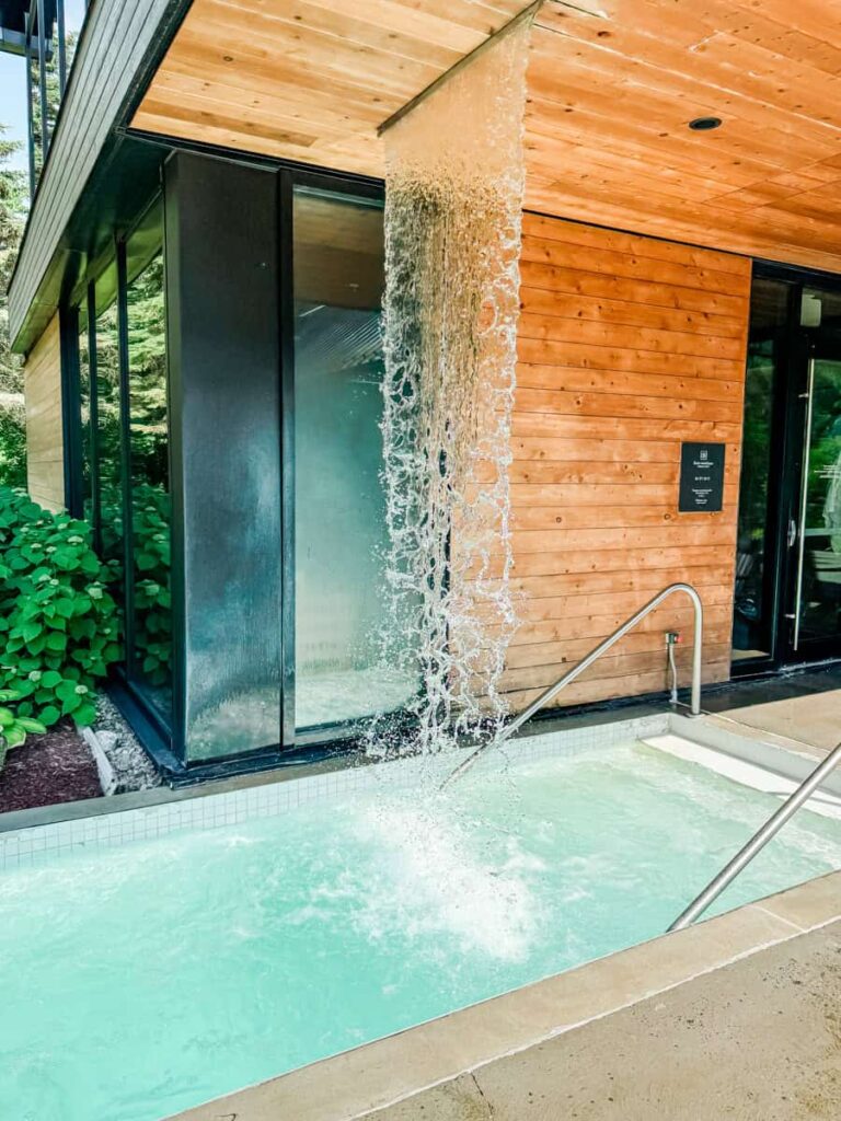 A stream of water cascades from a wooden roof into a turquoise spa pool at Strøm Spa Nordique, surrounded by glass walls and greenery.