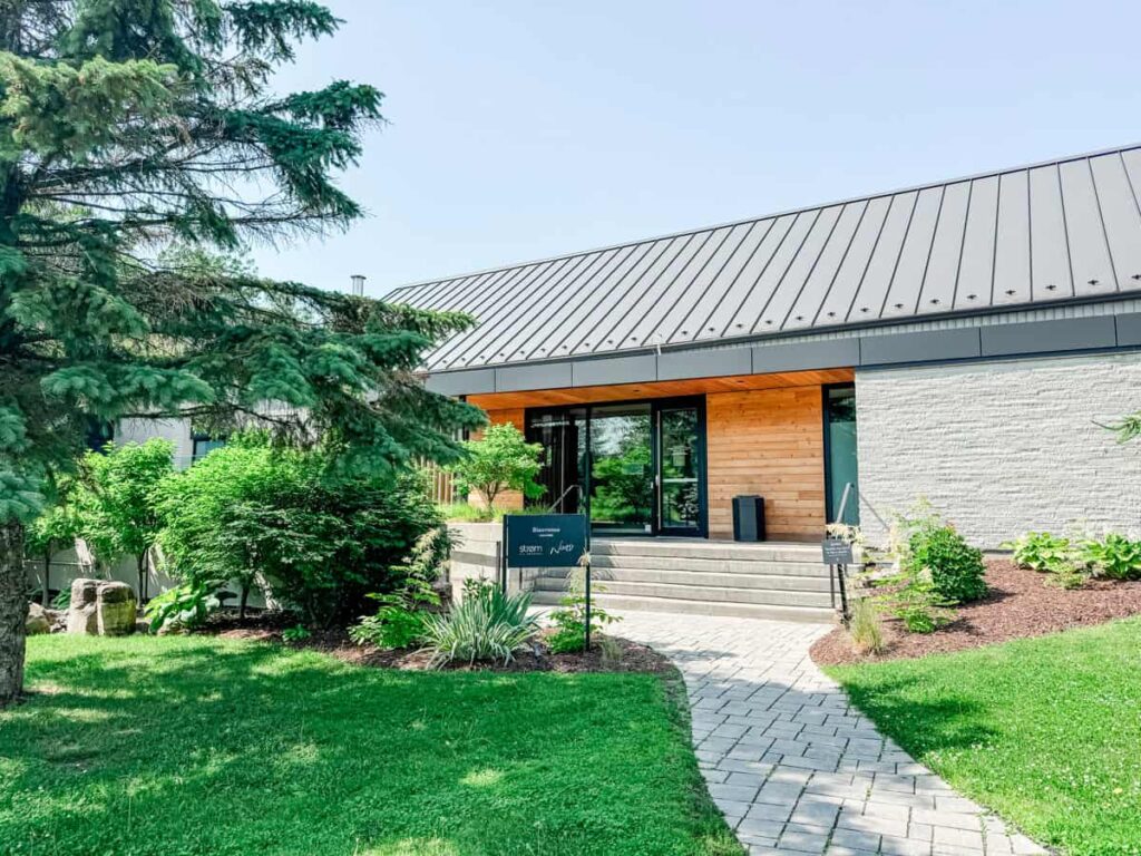 Exterior of Nord restaurant at Strom Spa Nordique, featuring modern wood and stone architecture surrounded by lush greenery and a curved stone walkway.