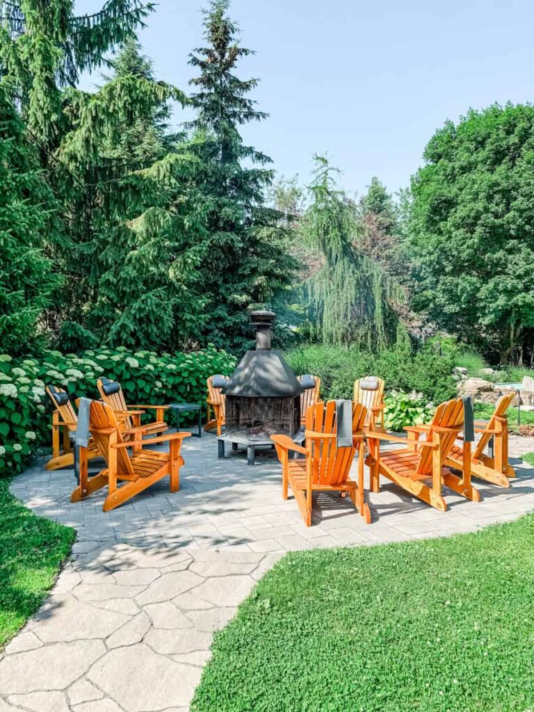 Circle of bright wooden Adirondack chairs with towels draped over them, gathered around a central outdoor fireplace in a lush garden setting.
