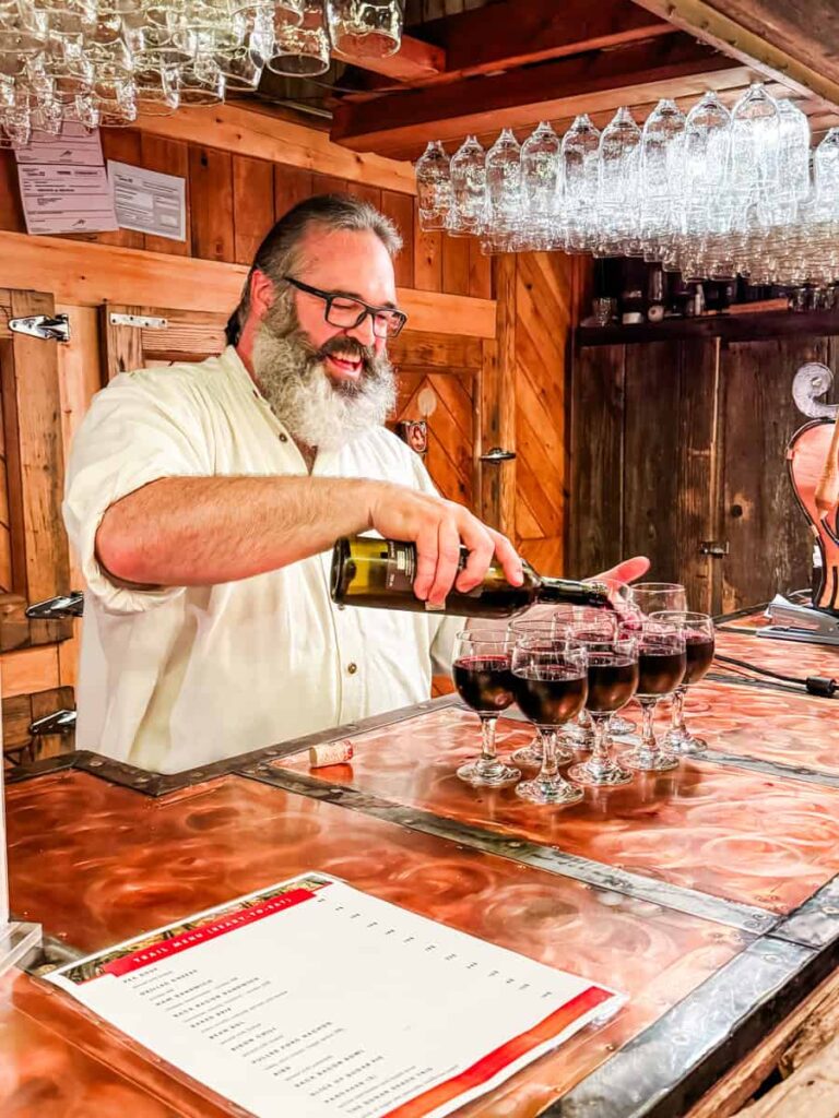 Stefan Faucher behind a copper bar pours Caribou red wine drinks into a row of glasses at Sucrerie de la Montagne, with upside-down mugs hanging above.