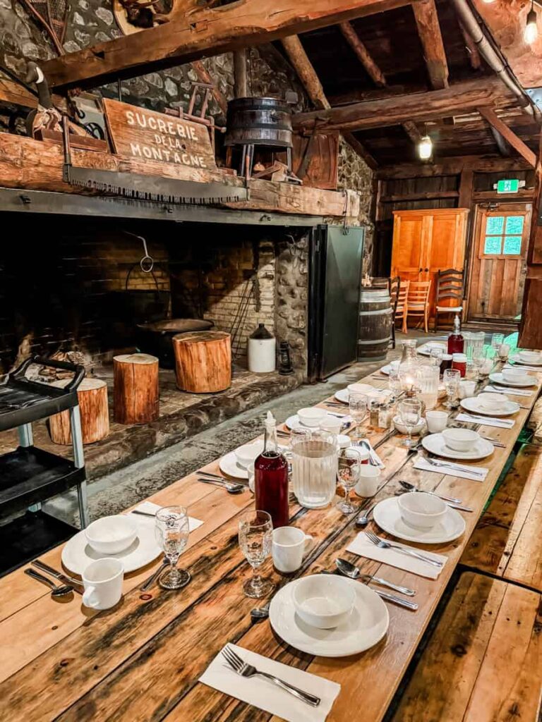 Long rustic dining table set with white dishes inside a log cabin-style dining room, featuring a stone fireplace and wooden beams.