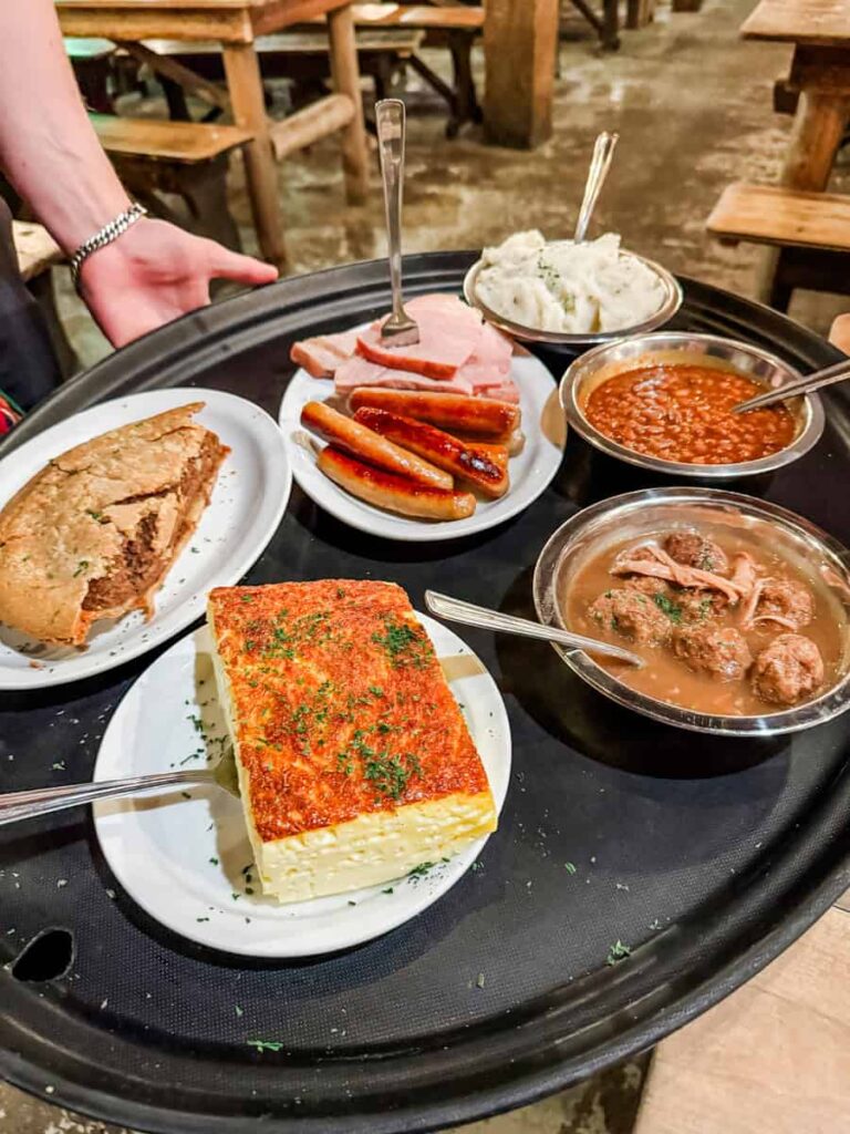 Tray of traditional Quebecois dishes including sausages, ham, mashed potatoes, meatballs in sauce, baked beans, omelette-style egg dish, and meat pie.