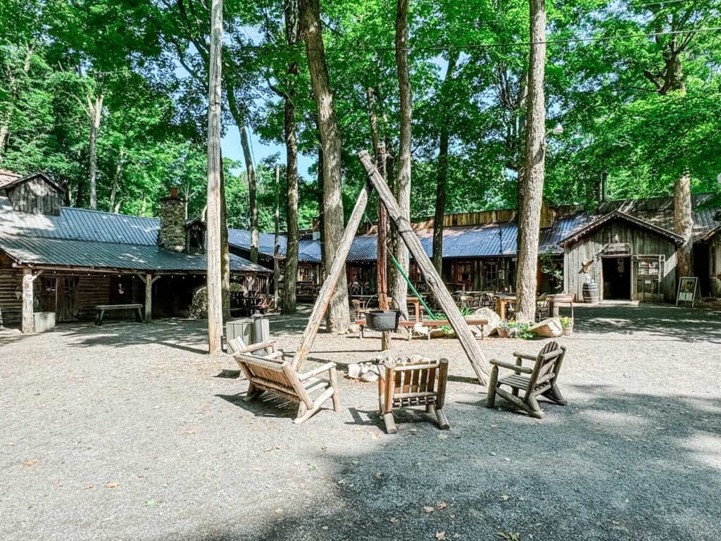 Outdoor seating area at Sucrerie de la Montagne, with rustic wooden chairs surrounding a tripod-hung cauldron over a fire pit, set among tall trees and historic log cabins.