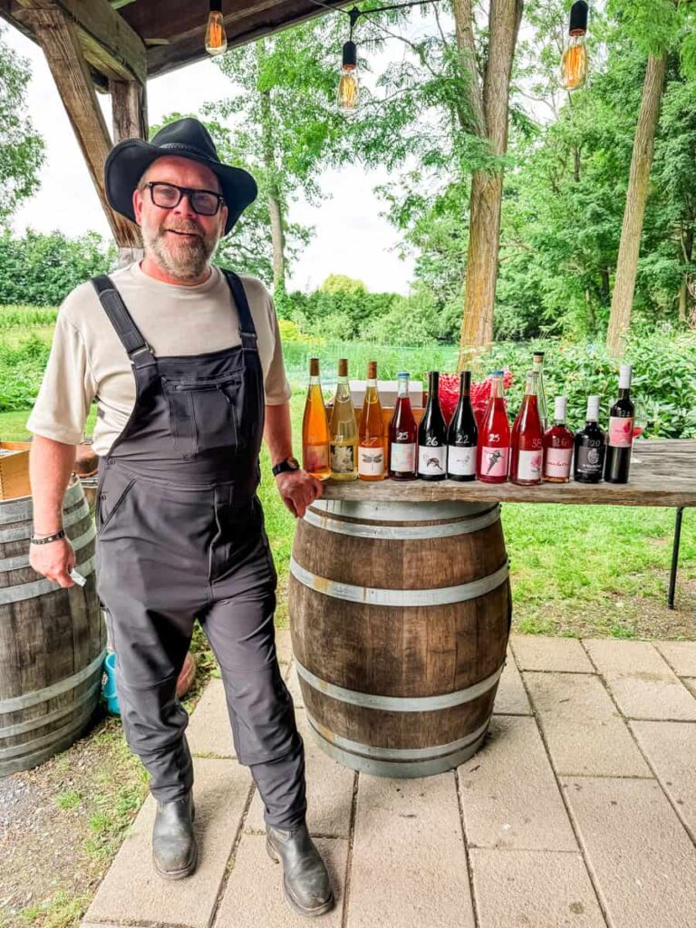 Pier Cousineau, co-owner and winemaker at Vignoble de l'Ardennais, standing beside a wine barrel showcasing an array of colorful wine bottles under a shaded pavilion.