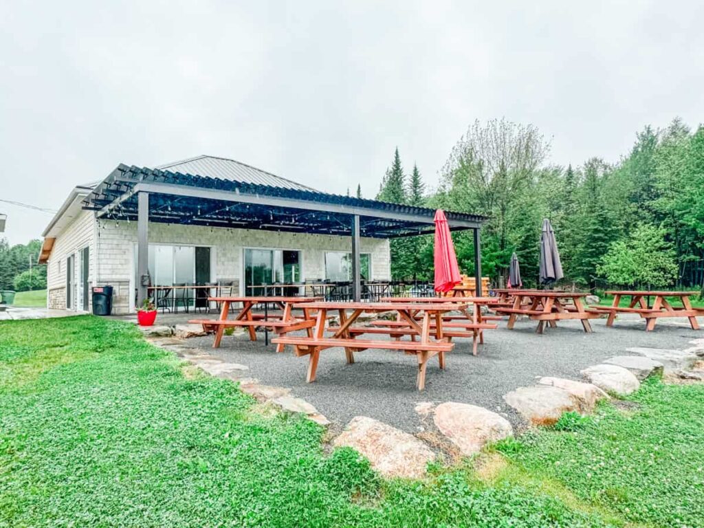 Spacious outdoor tasting patio at Vignoble Les Côtes du Gavet, with wooden picnic tables and umbrellas beside a modern stone building, surrounded by forest.