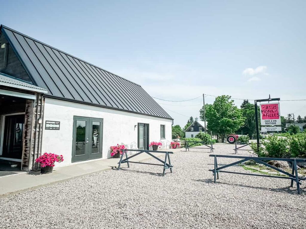 Exterior of Cassis Monna & Filles, featuring a modern white building with black trim, bright pink flowers in planters, and a sign for the boutique and wine cellar under a clear blue sky.