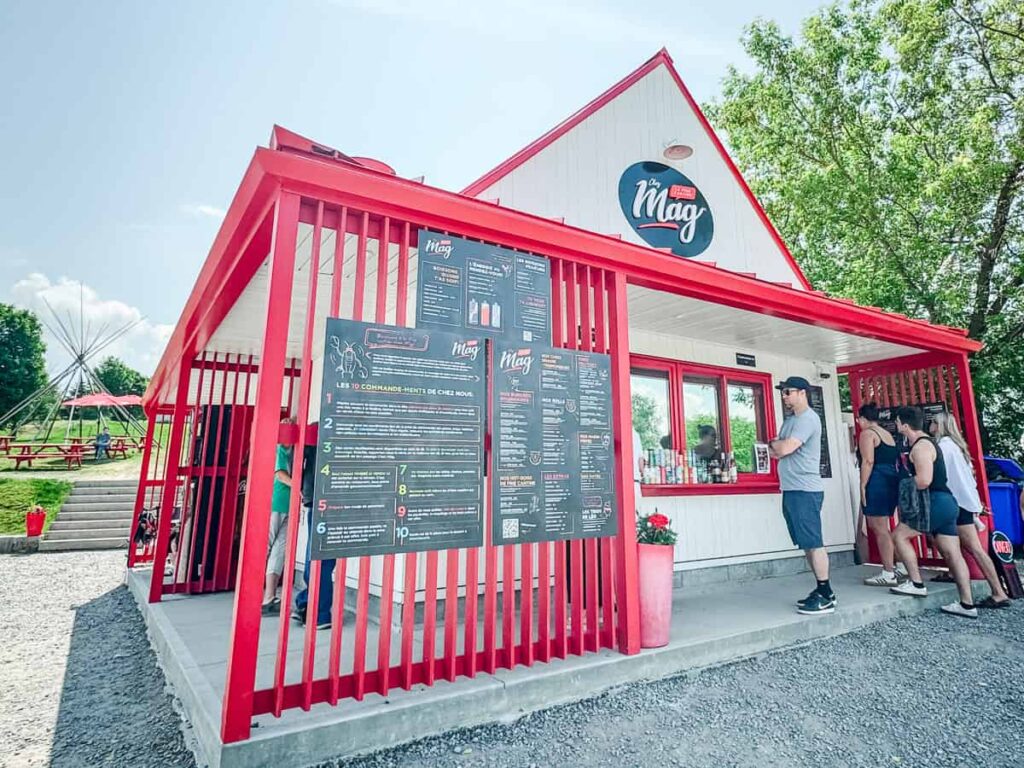 The front of Chez Mag restaurant with a red-and-white exterior, outdoor menu boards, and a line of customers waiting to order.