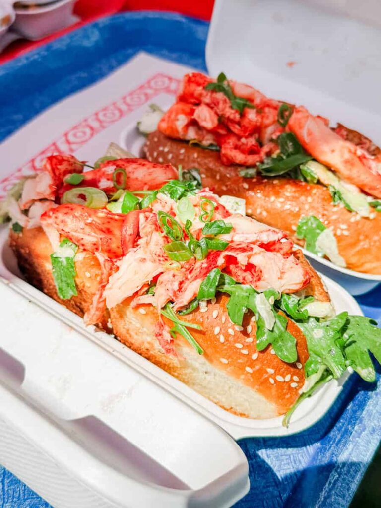 Two toasted sesame buns filled with lobster meat and fresh greens, served in a takeout container from Chez Mag.