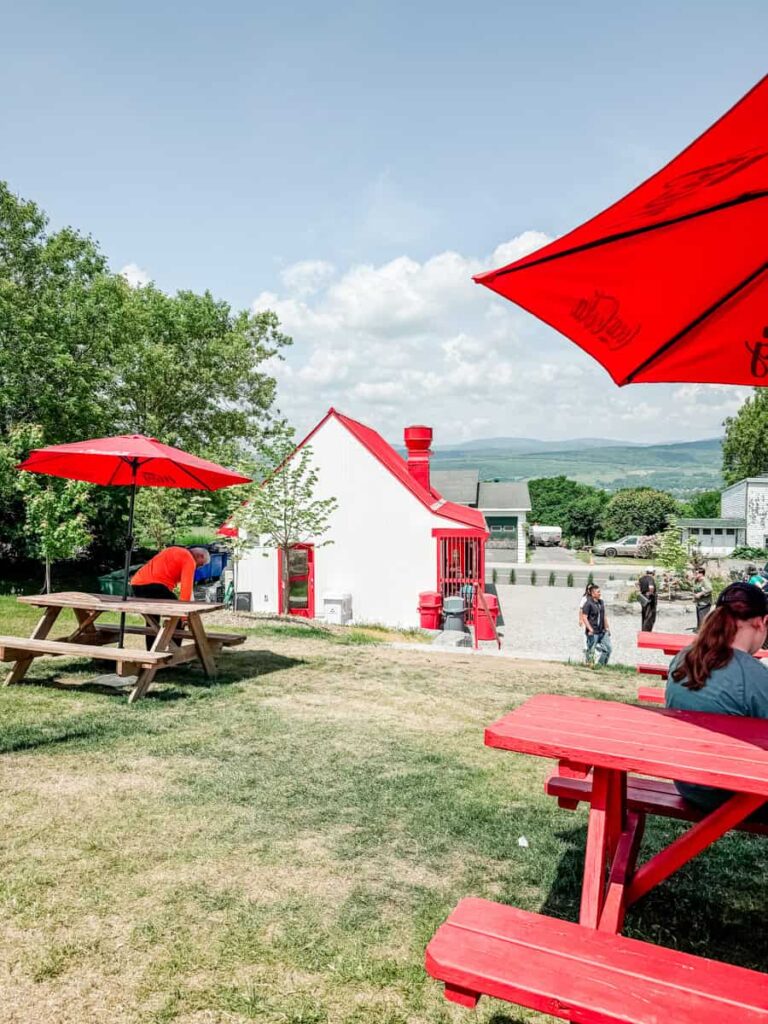 Outdoor seating area at Chez Mag with red picnic tables and umbrellas, overlooking a scenic rural landscape with a small white building in the background.