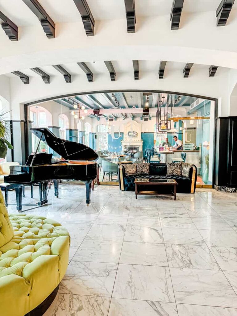 Elegant marble-floored lobby at Hotel Clarendon featuring a grand piano, plush green seating, and a view into the restaurant through glass walls.