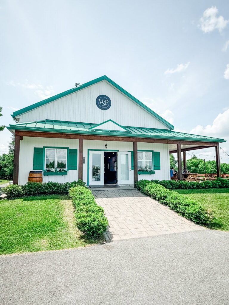 The charming tasting room at Vignoble Ste-Pétronille, featuring green shutters, a green metal roof, and manicured hedges lining the walkway.