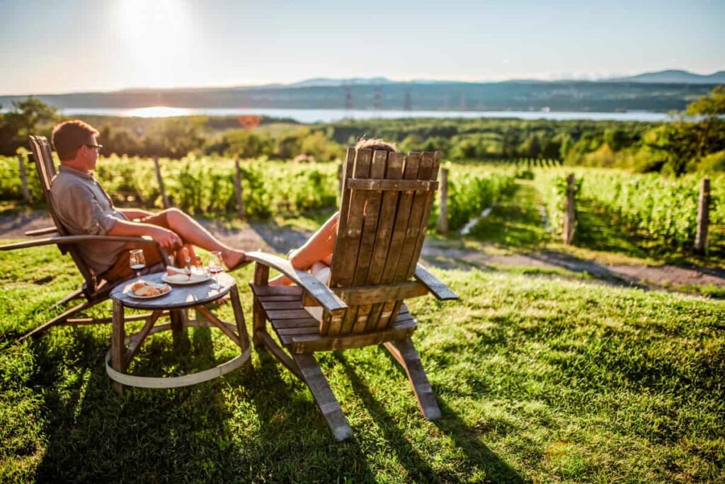 A couple relaxes in wooden chairs at Vignoble de l’Isle de Bacchus, sipping wine with vineyard rows and the St. Lawrence River glowing in the sunset.