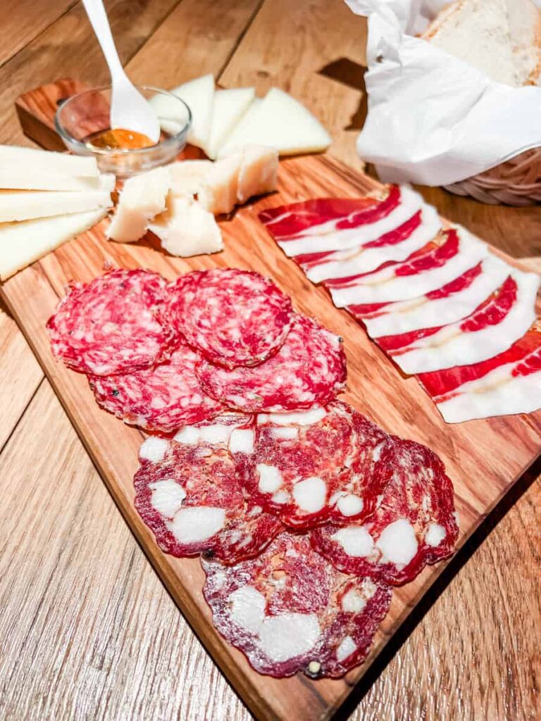 Assorted cured meats, cheeses, and a small dish of wine jelly on a wooden cheese board, with bread in a basket on a rustic wooden table.