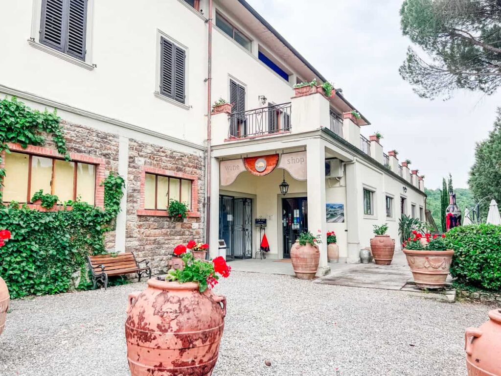 Castello Monterinaldi entrance with potted plants, outdoor seating, and greenery, surrounded by a rustic building with black shutters and a balcony, under a cloudy sky.