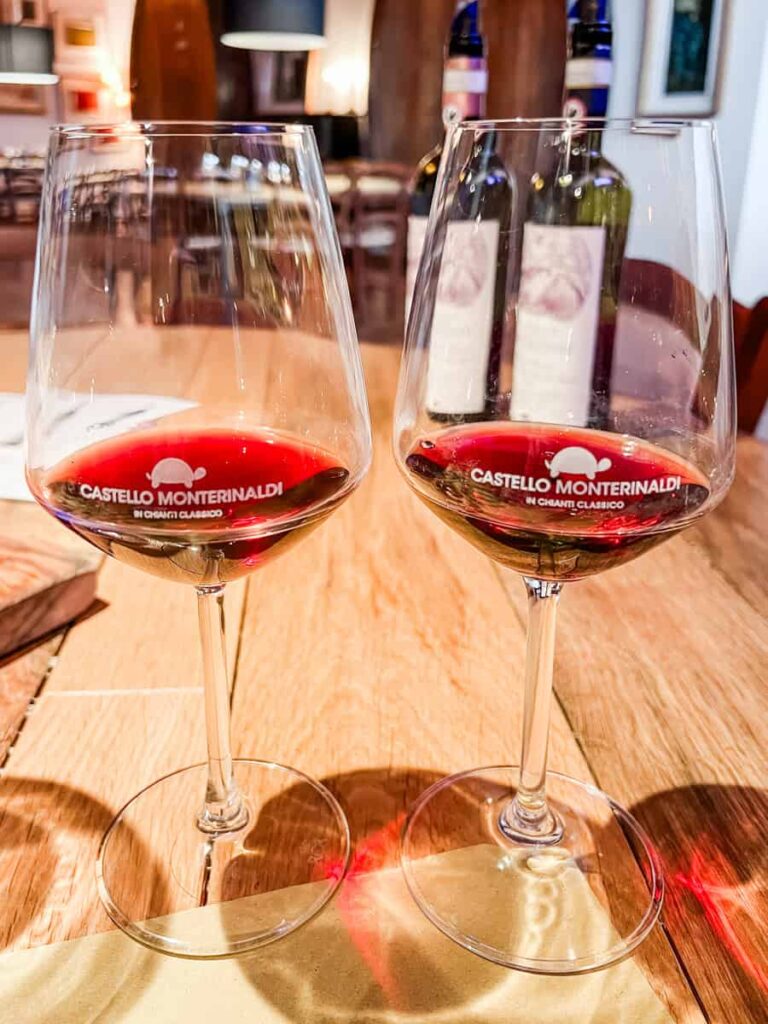 Two glasses of red wine on a wooden table with wine bottles in the background, showcasing a wine tasting experience at Castello Monternaldo.