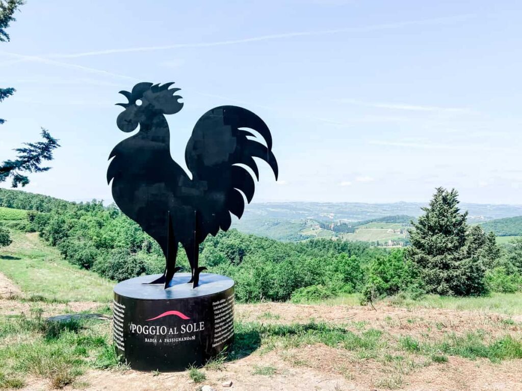 A black metal sculpture of a rooster standing on a circular base with scenic green hills and trees in the background on a sunny day.