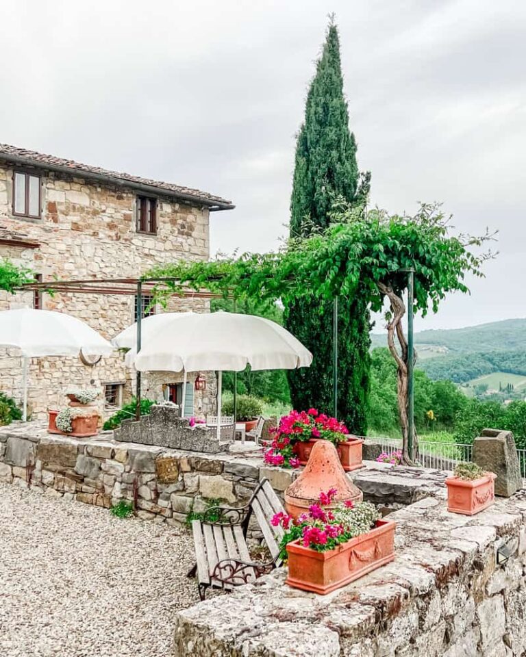 Stone tasting room at Il Barlettaio, a Chianti winery, with outdoor patio, pink flowers, white umbrellas, and rolling vineyards and hills in the background.