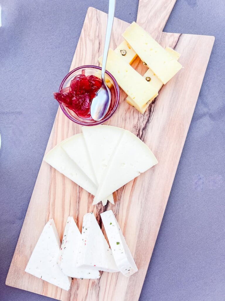 Assorted cheese platter with wedges of cheese, a small bowl of red fruit preserves, and a spoon on a wooden serving board.