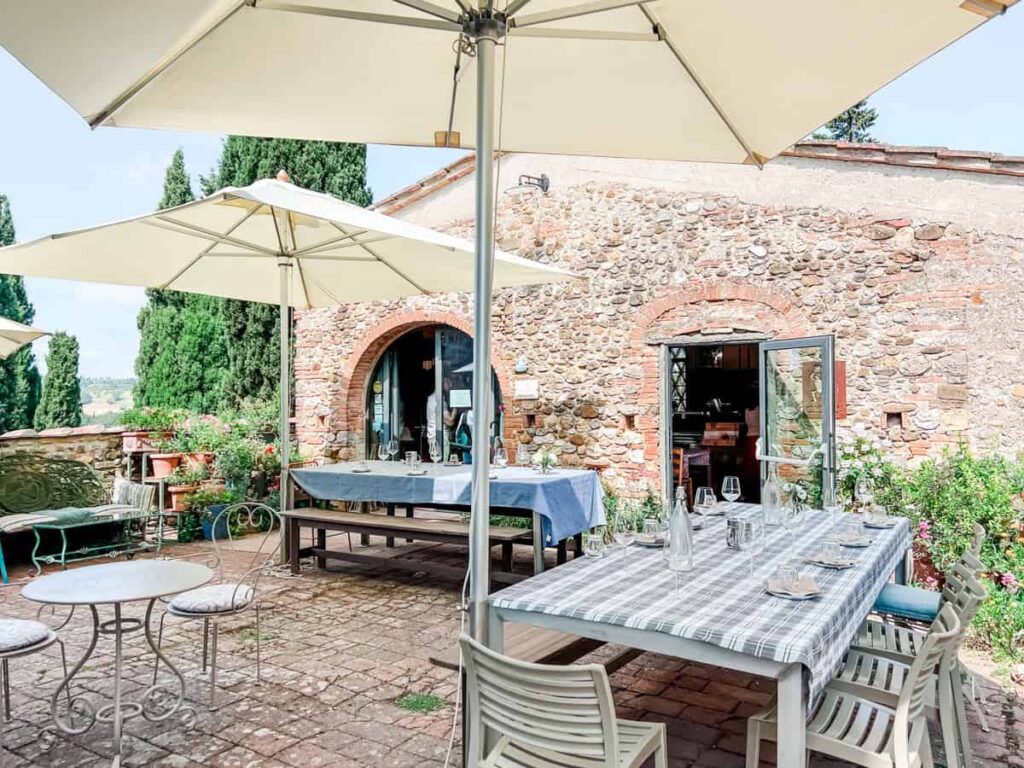 Rustic outdoor wine tasting area with a brick building, white umbrellas, and a lush garden setting. Elegant table arrangements with glassware and checkered tablecloths create a charming atmosphere.