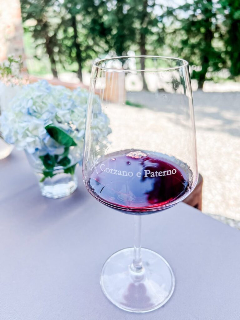 A glass of red wine on a table at Corzano e Paterno with a blurred bouquet of white and pale blue flowers in the background outside with trees.