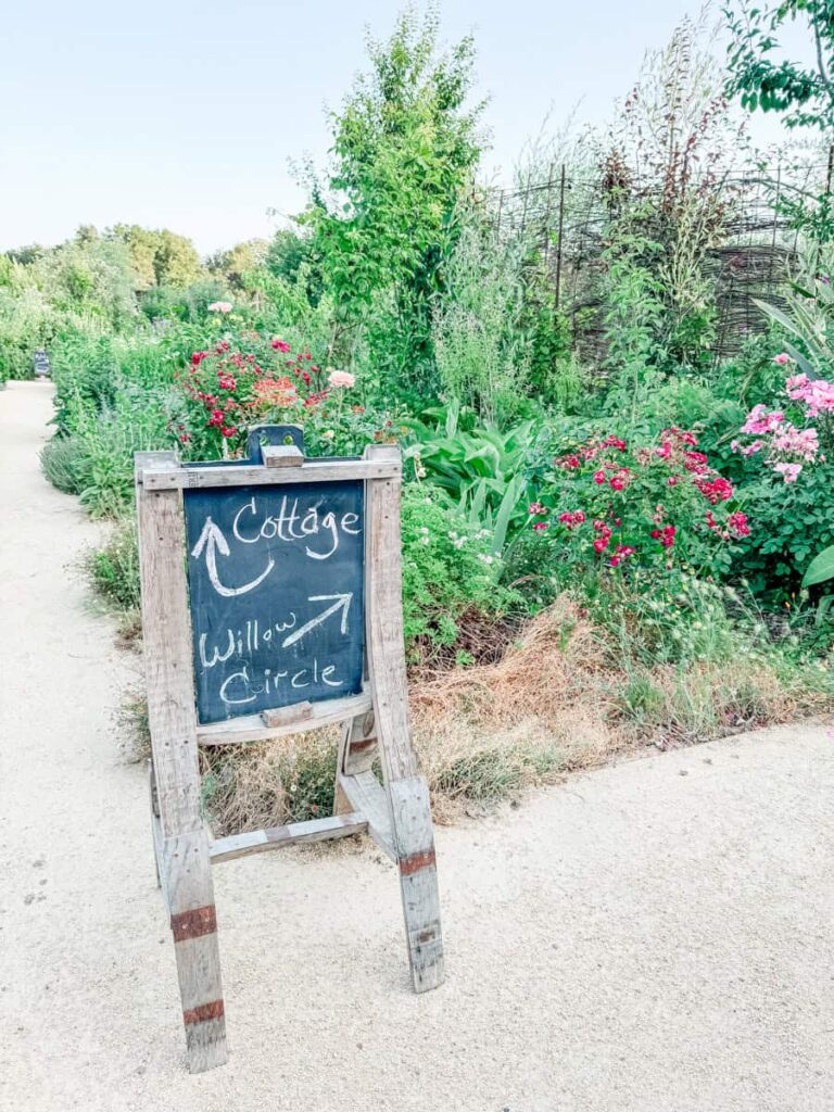 Handwritten chalkboard sign at DaVero pointing the way to the cottage and Willow Circle, set beside a vibrant garden path with blooming flowers and greenery.