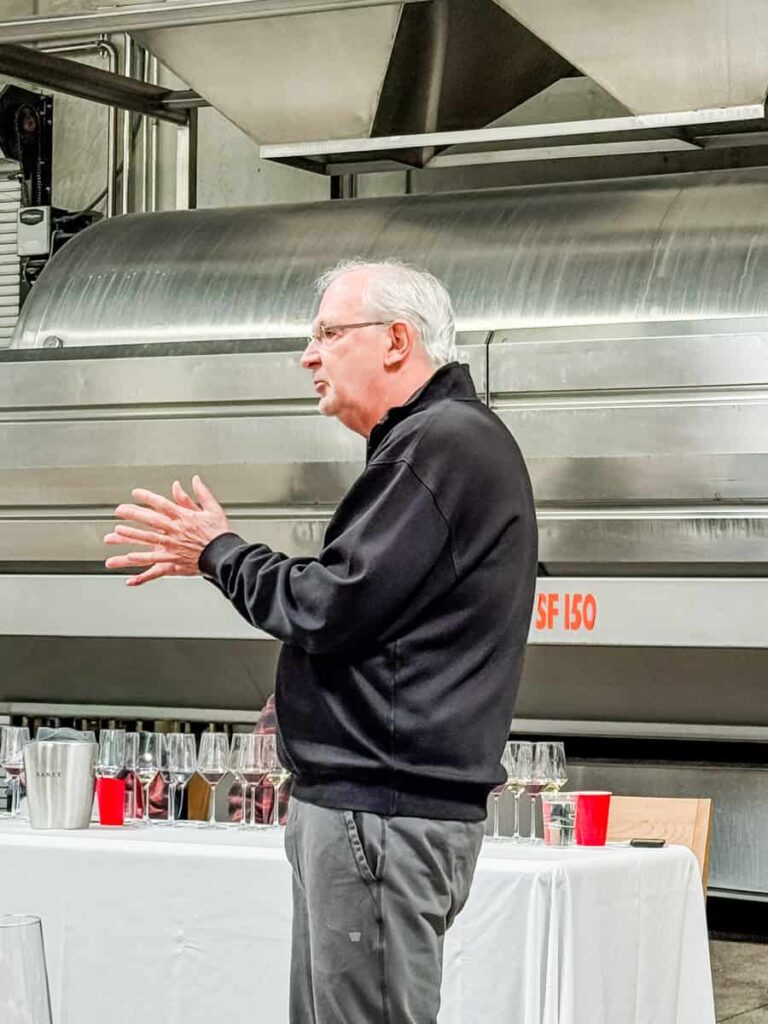 David Ramey of Ramey Cellars giving a tasting presentation next to a stainless steel wine press and a table with glasses and tasting accessories on a white tablecloth.