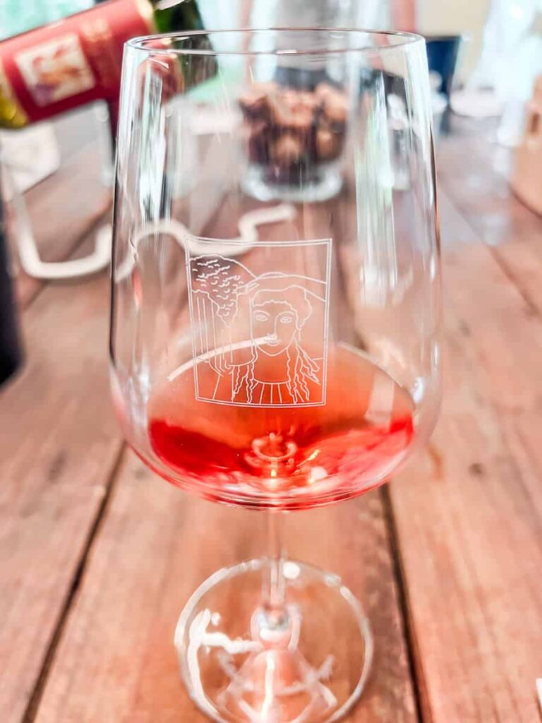 A glass of Fattoria di Montemaggio rosé wine with an etched illustration of a woman's face on the glass, placed on a rustic wooden table with blurred wine bottles in the background.