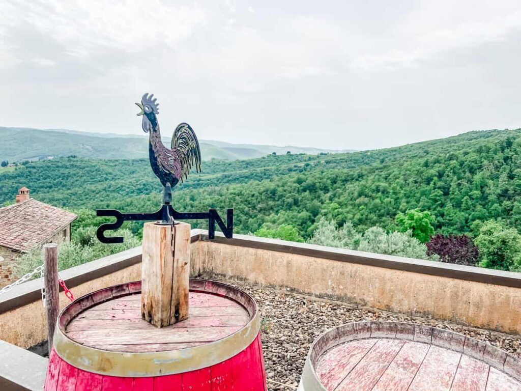 Colorful rooster weather vane atop wine barrels overlooking a green landscape at Fattoria di Montemaggio in the Chianti region.
