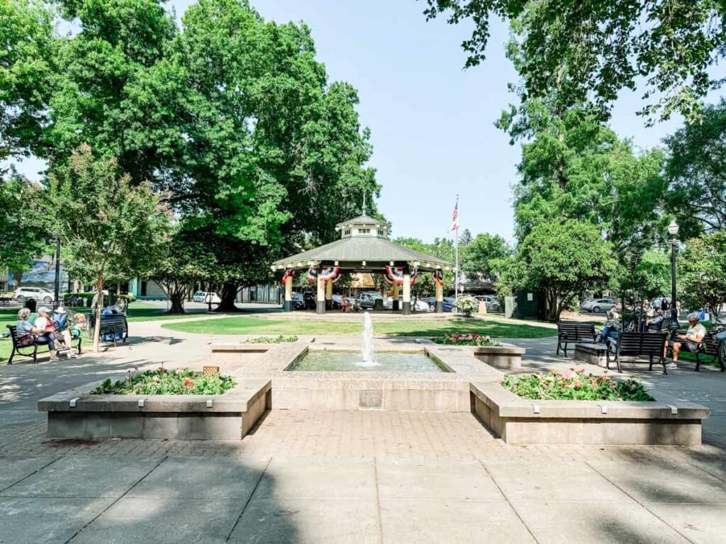 Healdsburg square with a central fountain surrounded by green trees, benches, and a pavilion decorated with red, white, and blue bunting, under a clear blue sky.
