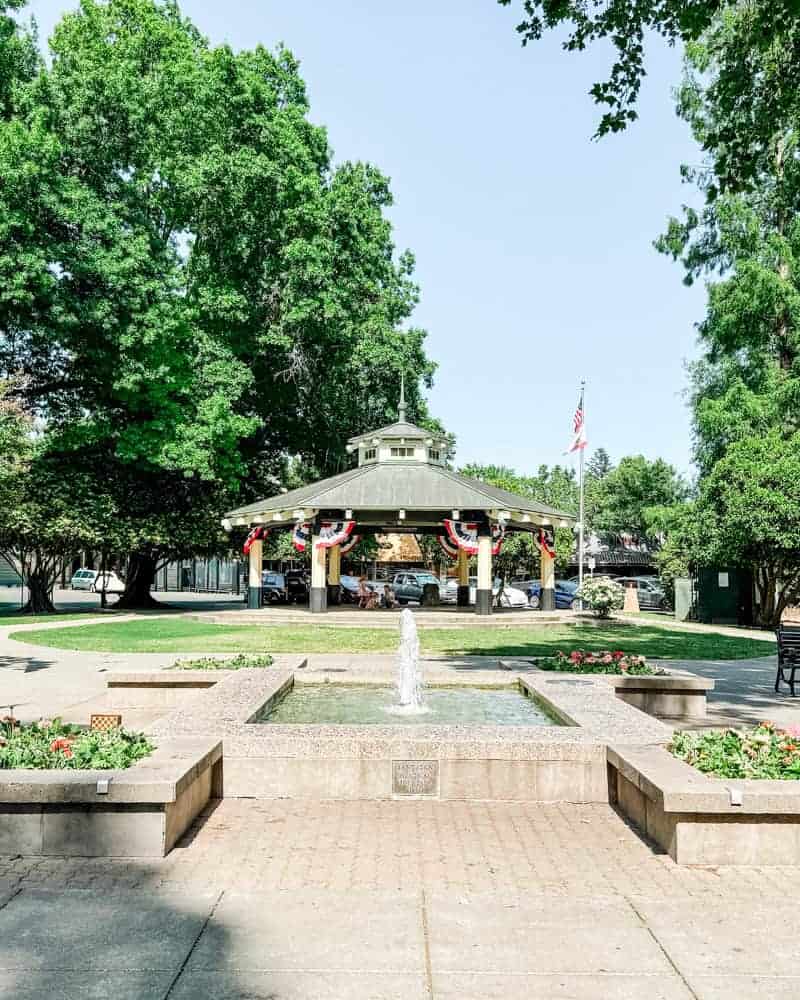 Healdsburg square with a central fountain surrounded by green trees, benches, and a pavilion decorated with red, white, and blue bunting, under a clear blue sky.