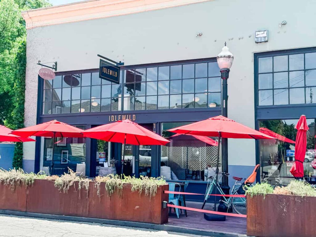 Outdoor patio and entrance of Idlewild's tasting room with large glass windows, red umbrellas shading seating, and decorative plants in planters.
