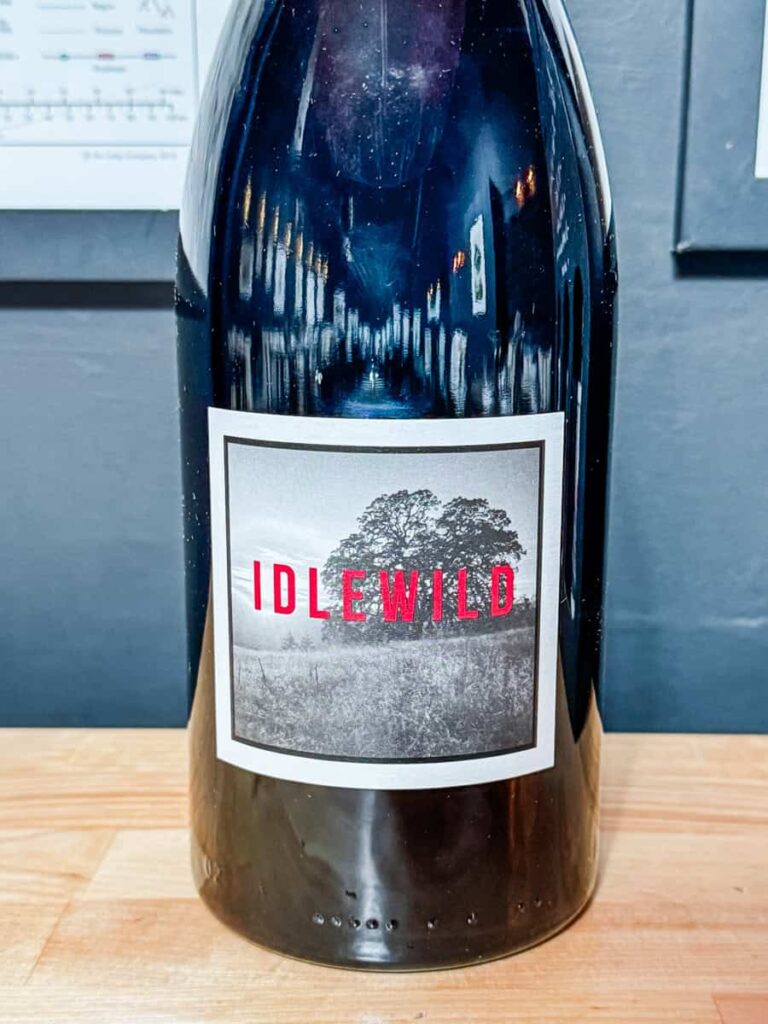 A dark glass wine bottle with a white label featuring a black and white illustration of a tree and background landscape, with the word "IDLEWILD" in bold red capital letters. The bottle is placed on a light wooden surface, with blurred dark background elements.